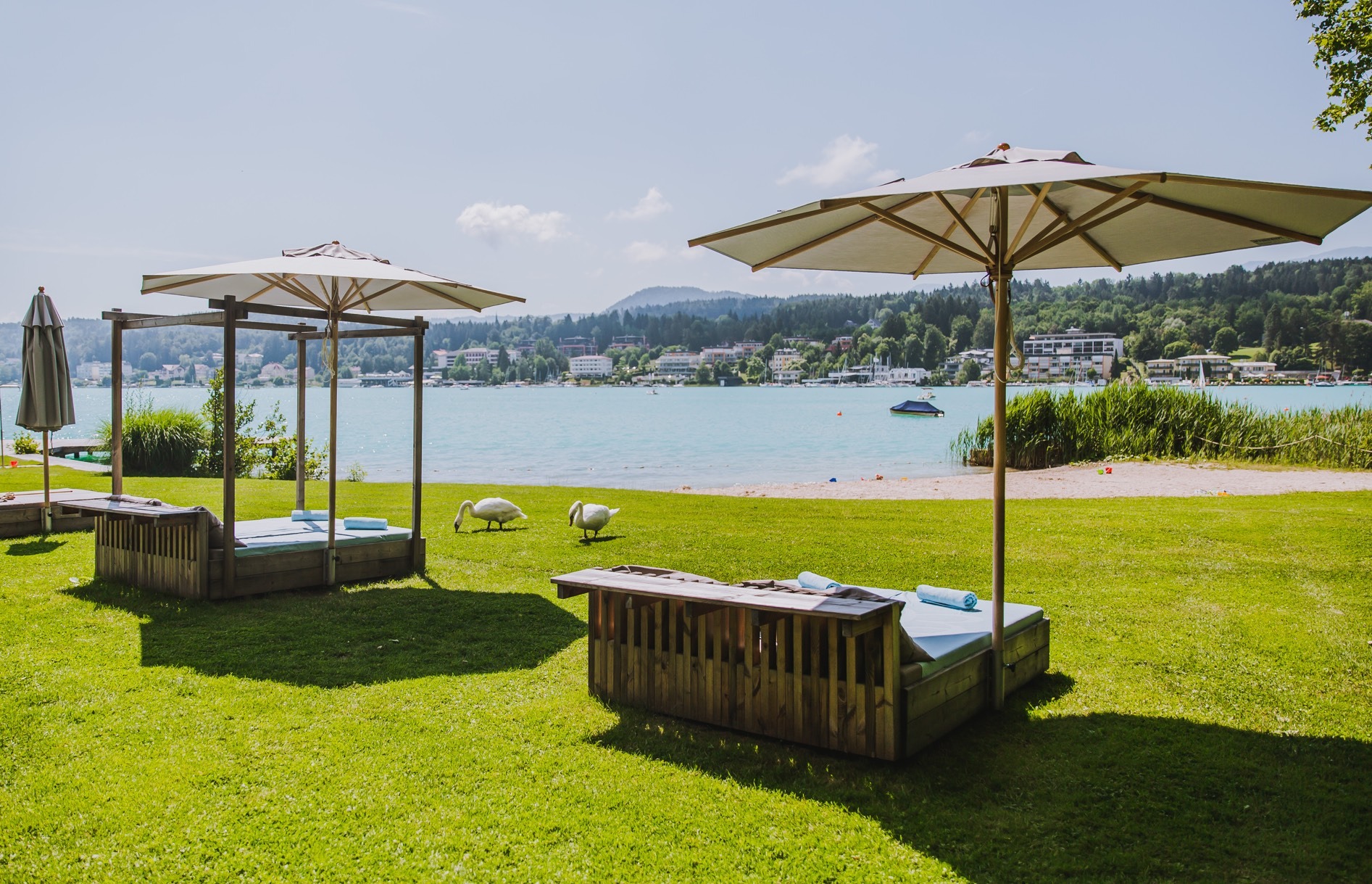 Hölzerne Daybeds mit Sonnenschirmen auf der Liegewiese, dahinter Strand und Wörthersee